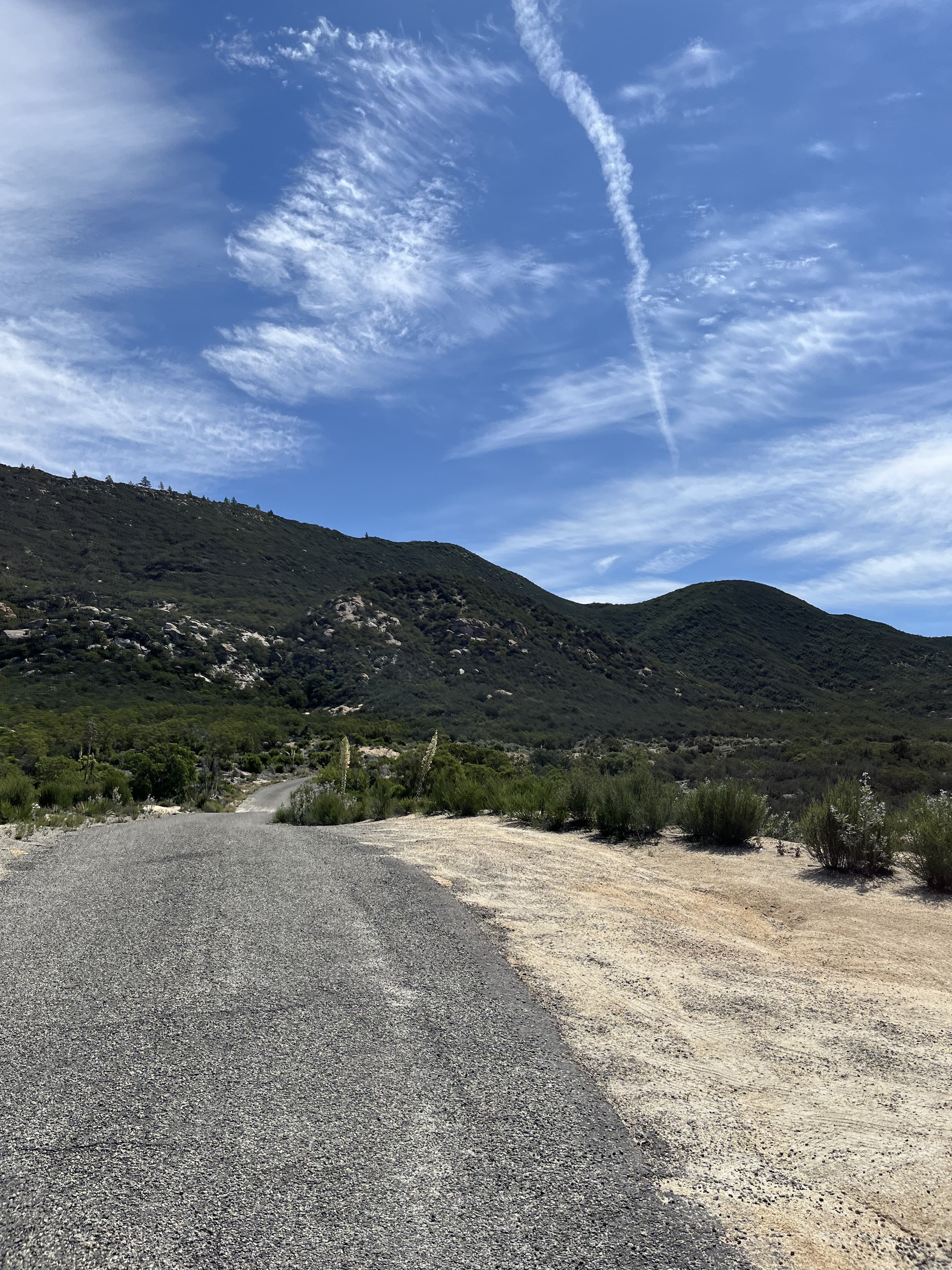 Dirt road descending through green mountains toward San Diego, with a jet contrail overhead